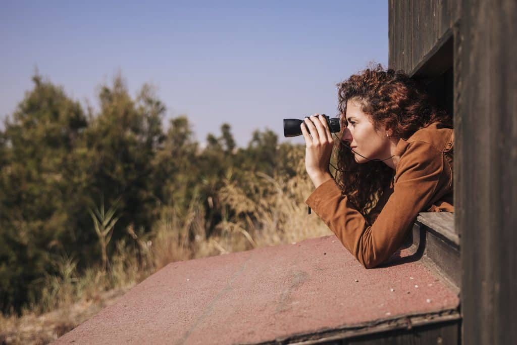 A redhead woman leaning out and peering through binoculars during the best time for bird watching in Kruger.