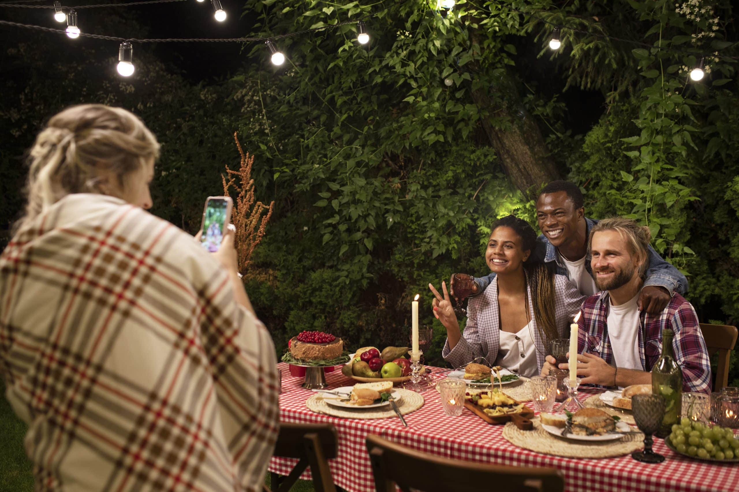 Friends pose for a photo at an outdoor dinner table under string lights, enjoying food and wine in a garden at night.